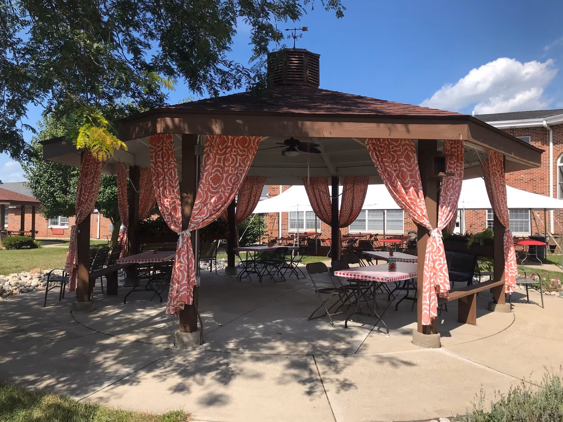 Open wooden gazebo with pink patterned curtains and folding tables and chairs set up in a courtyard in front of a brick building.