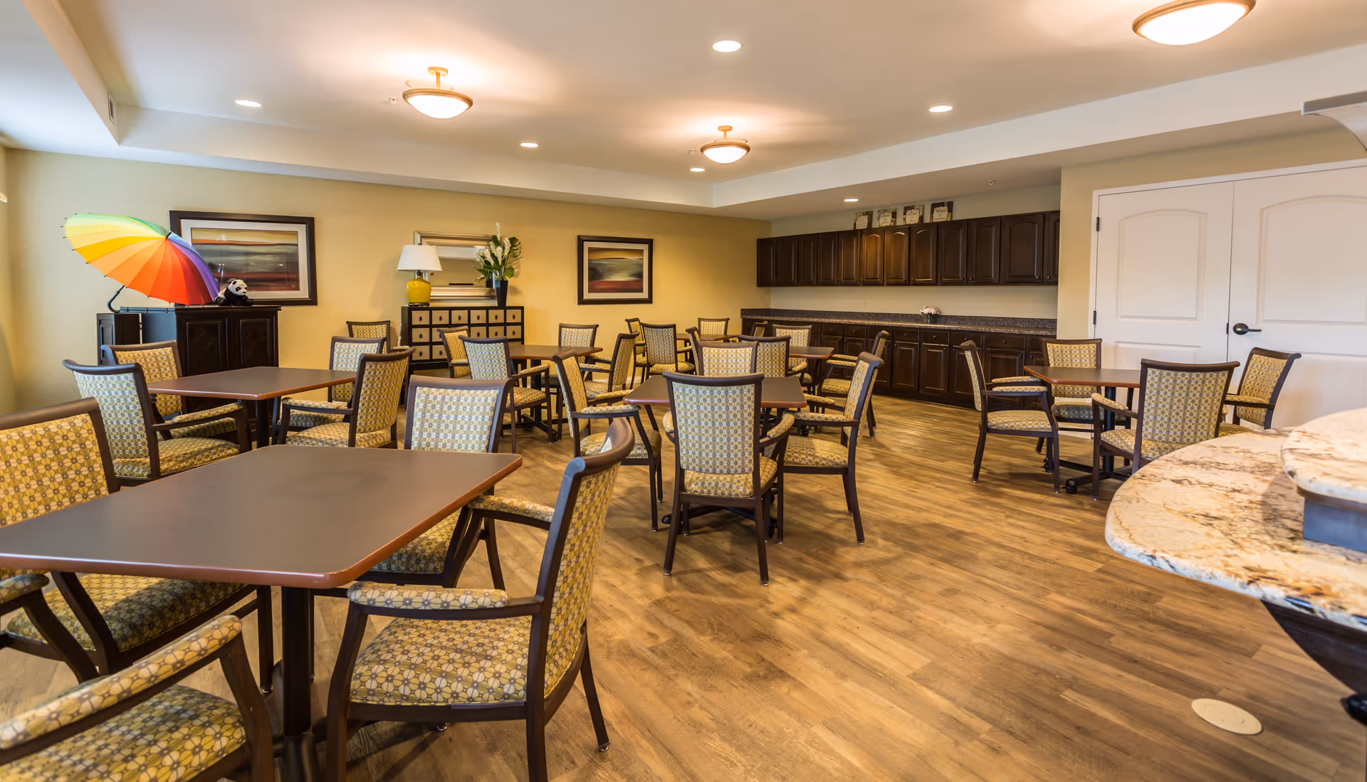 A spacious dining room with multiple square tables and cushioned chairs arranged neatly on a wooden floor. The walls are painted light yellow and decorated with framed artwork. There is a cabinet with a colorful umbrella and a small stuffed panda on top. The room has ceiling lights and a counter with dark wooden cabinets along one wall.