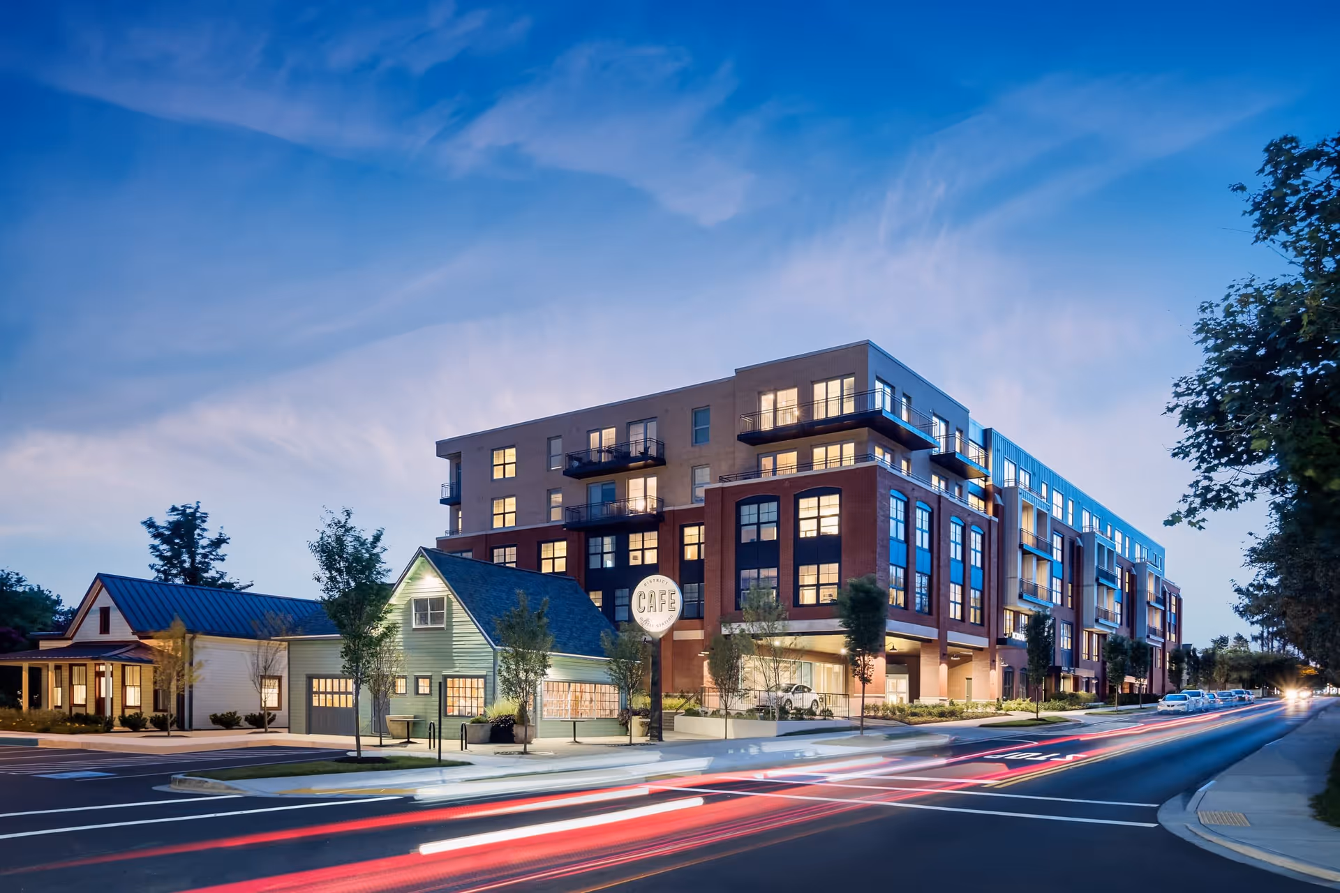 Evening view of a modern multi-story building with large windows and balconies next to smaller houses, with light trails from passing cars on the street in front.