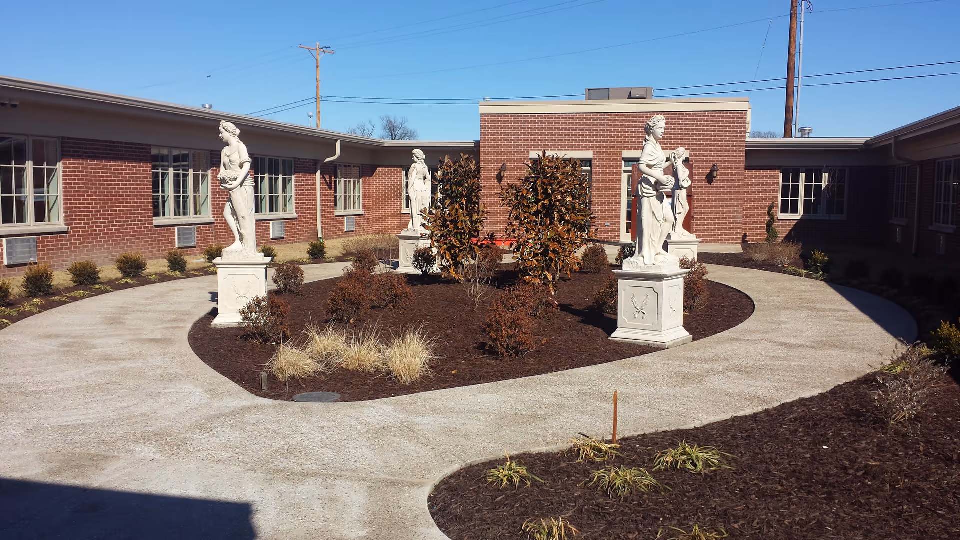 Circular outdoor courtyard with a paved path, landscaped beds and several classical statues in front of a low brick building.