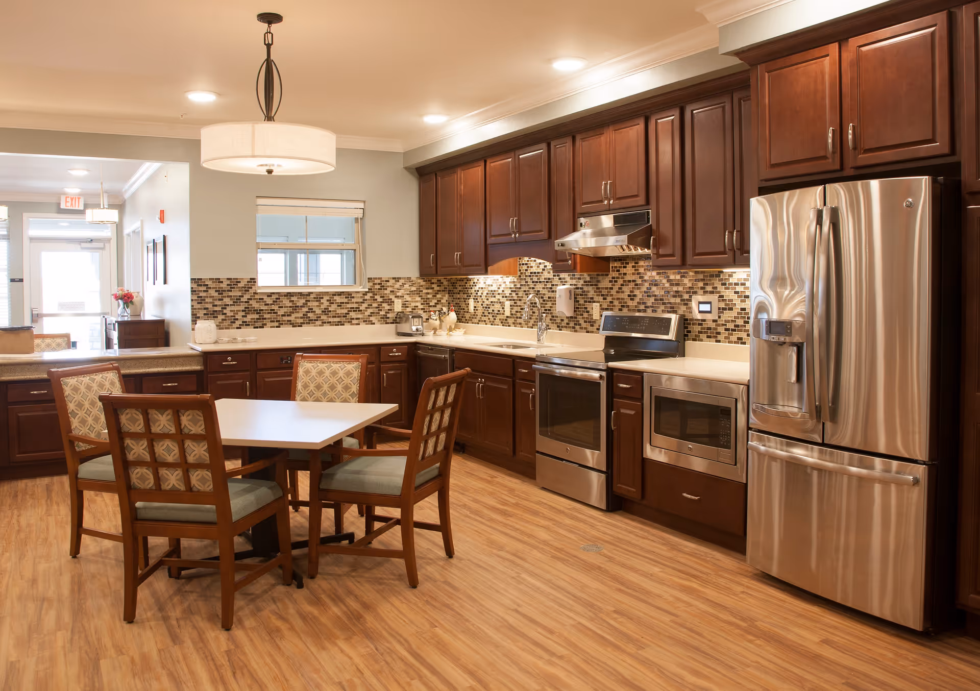 A spacious kitchen area with wooden cabinets, a stainless steel refrigerator, oven, microwave, and a tiled backsplash. In the center, there is a square dining table with four cushioned chairs. The room has wood flooring and is well-lit with ceiling lights and a hanging lamp above the table.