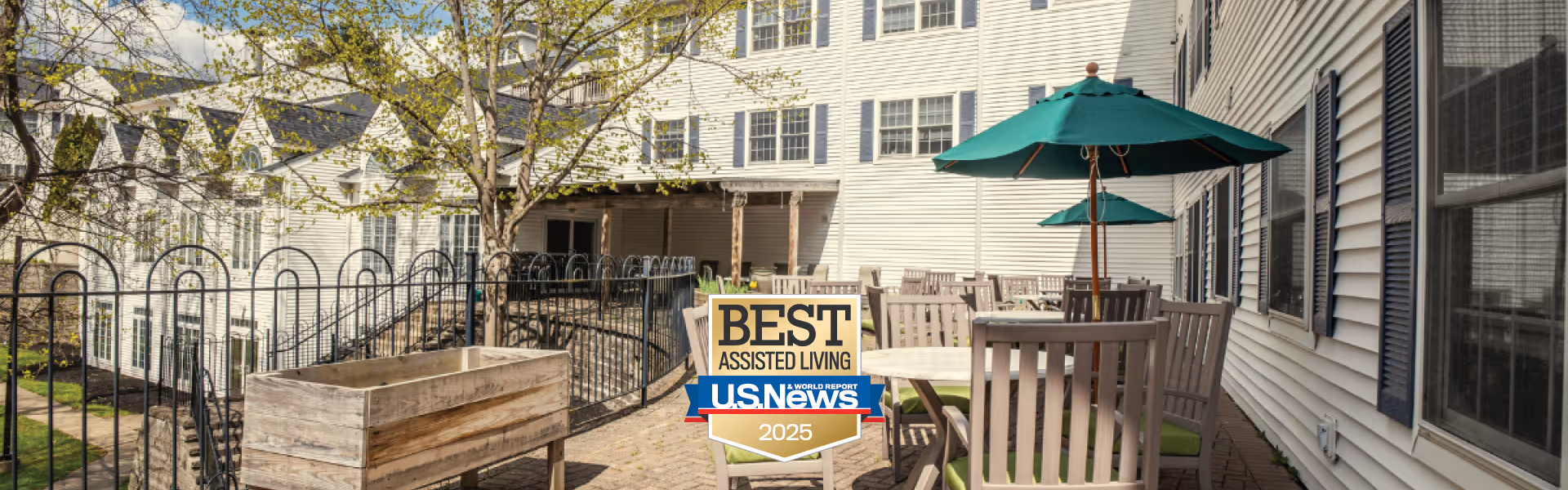 Outdoor patio area at Middlebrook Farms at Trumbull with wooden tables and chairs, green umbrellas, and a raised garden bed, adjacent to a white multi-story building with multiple windows.