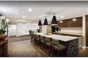 Modern kitchen and dining area in a retirement facility with a long island counter featuring stone accents and multiple bar stools. The space is well-lit with recessed lighting and three large pendant lights hanging above the island. There are dark wood cabinets and a light-colored countertop, with a small plant centerpiece on the island. The floor is wooden, and the background shows a hallway and a door.