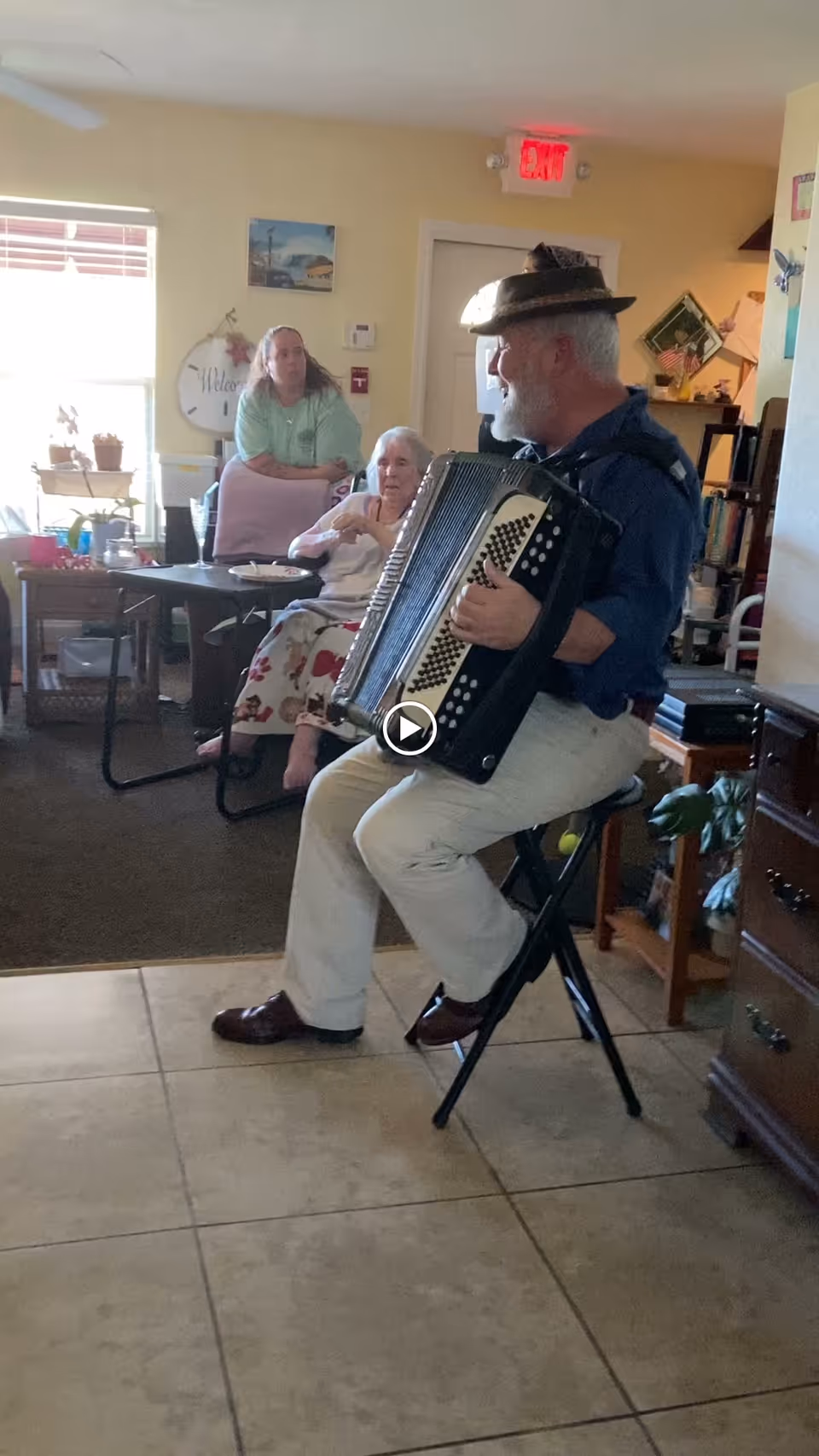 An elderly man wearing a hat is sitting on a folding stool playing an accordion in a cozy room. Two women are seated nearby, one on a chair and the other on a couch, attentively watching the performance. The room has tiled flooring near the man and carpeted flooring where the women sit, with various furniture and decorations visible in the background.
