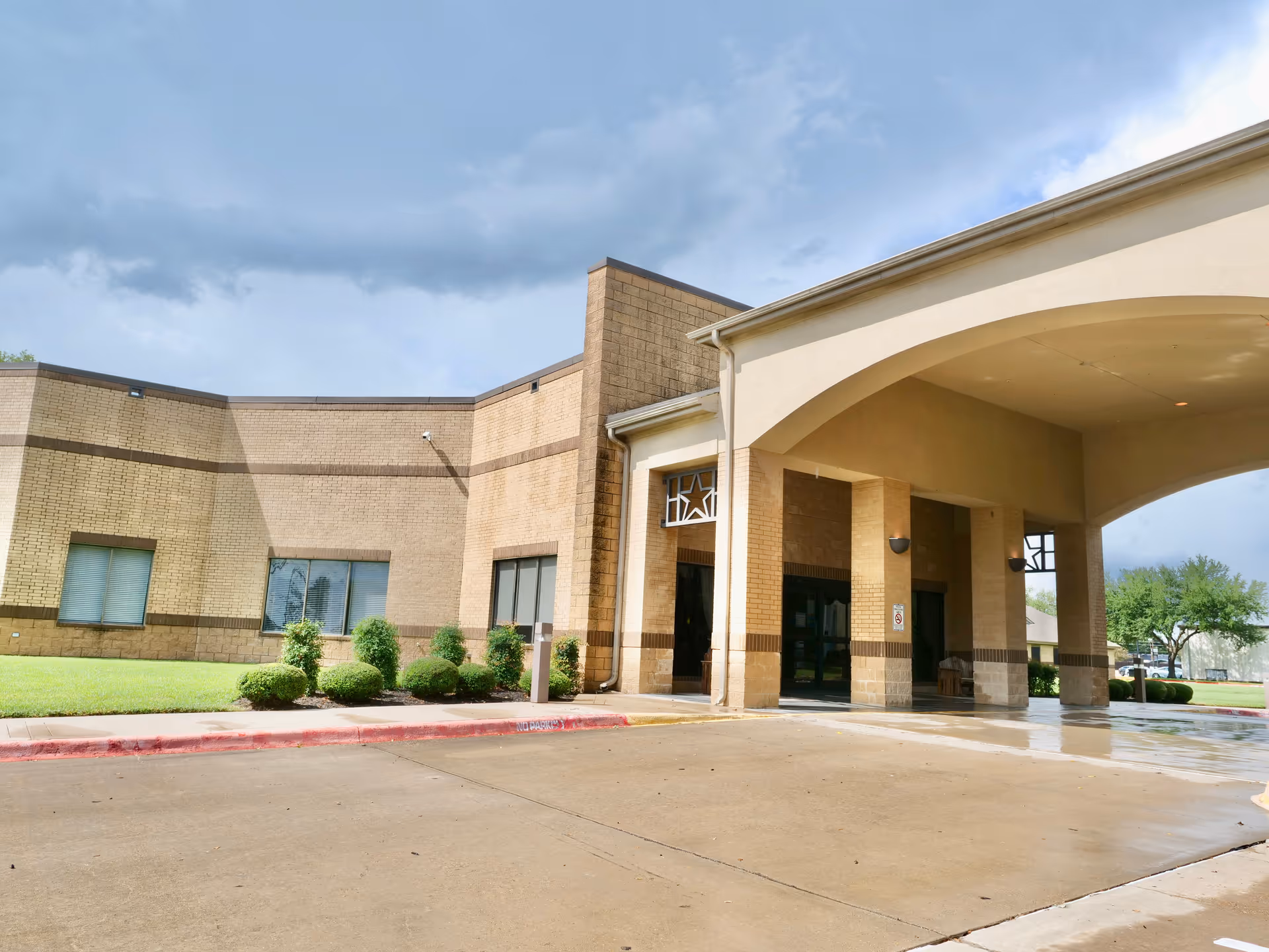 Exterior view of Twin Pines Nursing & Rehab Center showing the entrance with a covered drop-off area, beige brick walls, windows, and neatly trimmed bushes under a cloudy sky.
