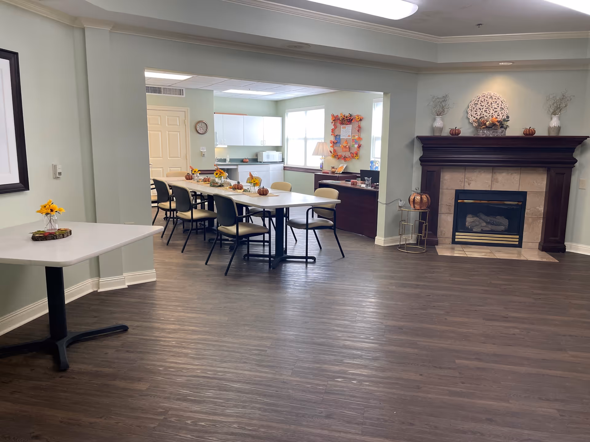 Interior view of a senior living facility common area with a fireplace on the right, decorated with vases and small pumpkins. In the background, there is a dining area with a long table surrounded by chairs, decorated with small fall-themed centerpieces. The walls are light green, and the floor is dark wood.