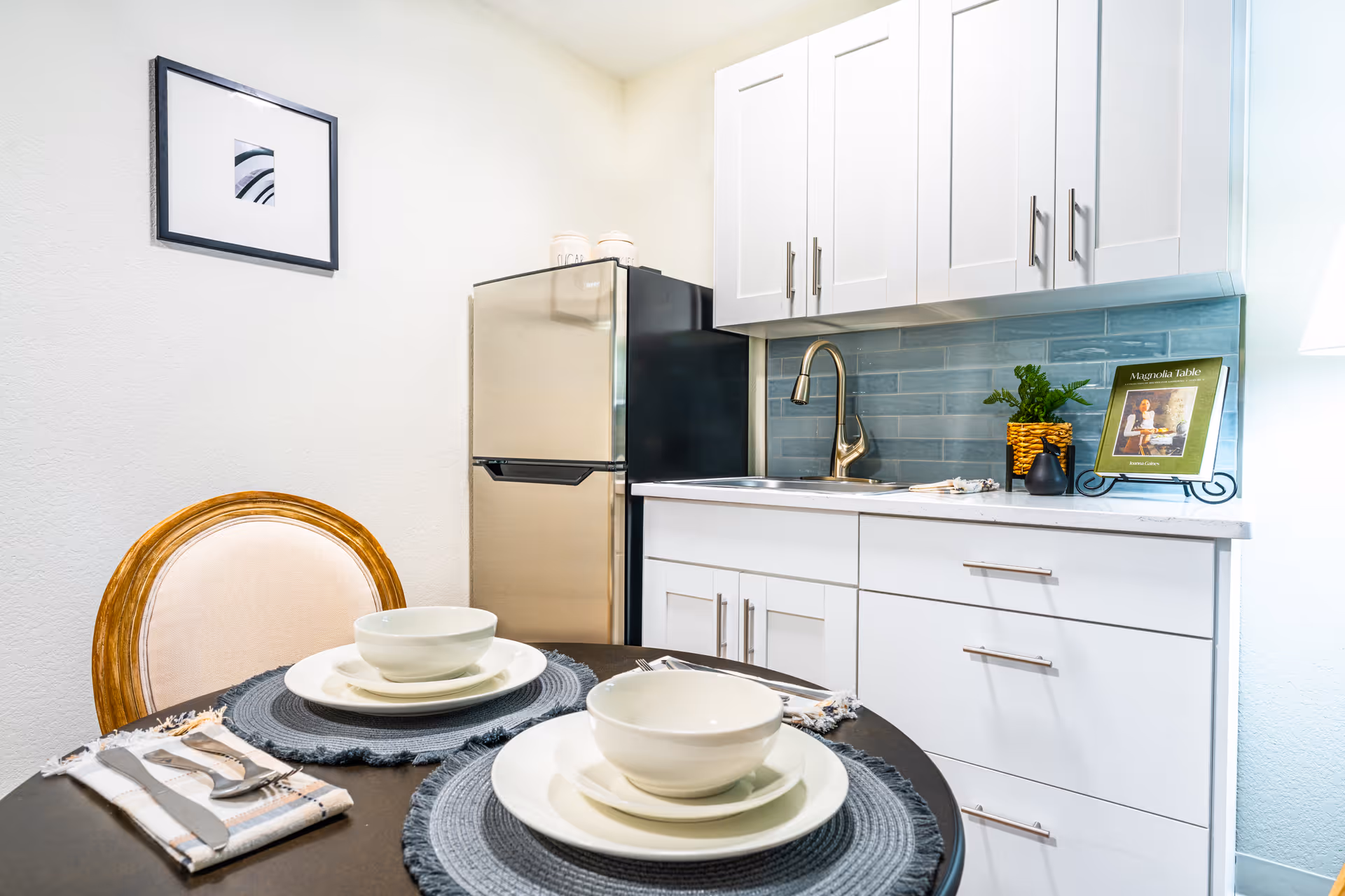 A small kitchen area with white cabinets, a stainless steel refrigerator, and a sink with a modern faucet. A round dining table is set with two place settings, each including a bowl, plate, and utensils on a gray placemat. There is a framed picture on the wall and a small plant and a cookbook on the kitchen counter.
