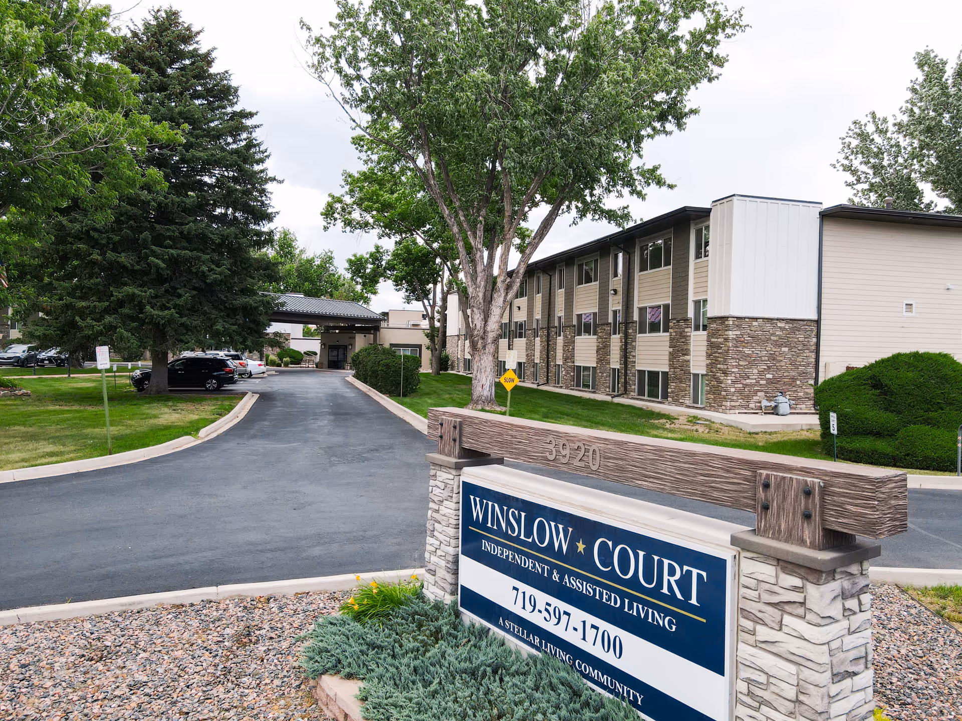 Exterior view of Winslow Court Assisted & Senior Living facility showing a two-story building with beige siding and stone accents, a driveway leading to the entrance, surrounded by green trees and grass, and a sign in the foreground with the facility name, phone number, and description.