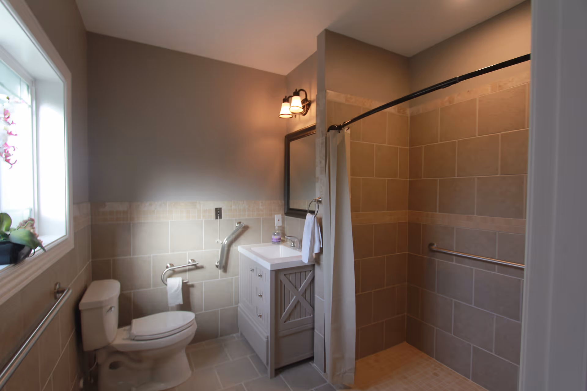 A clean and modern bathroom with beige tiled walls and floor. The bathroom features a white toilet with grab bars, a white vanity with a sink and mirror above it, a towel ring with a white towel, and a walk-in shower with a curved shower rod and curtain. There is a window with a plant on the windowsill allowing natural light to enter.