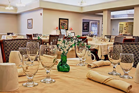 A dining room with tables covered in beige tablecloths, set with multiple empty glasses, rolled beige napkins, and a small green vase with white flowers as a centerpiece. The room has upholstered chairs and framed artwork on the walls, with soft lighting from ceiling fixtures.