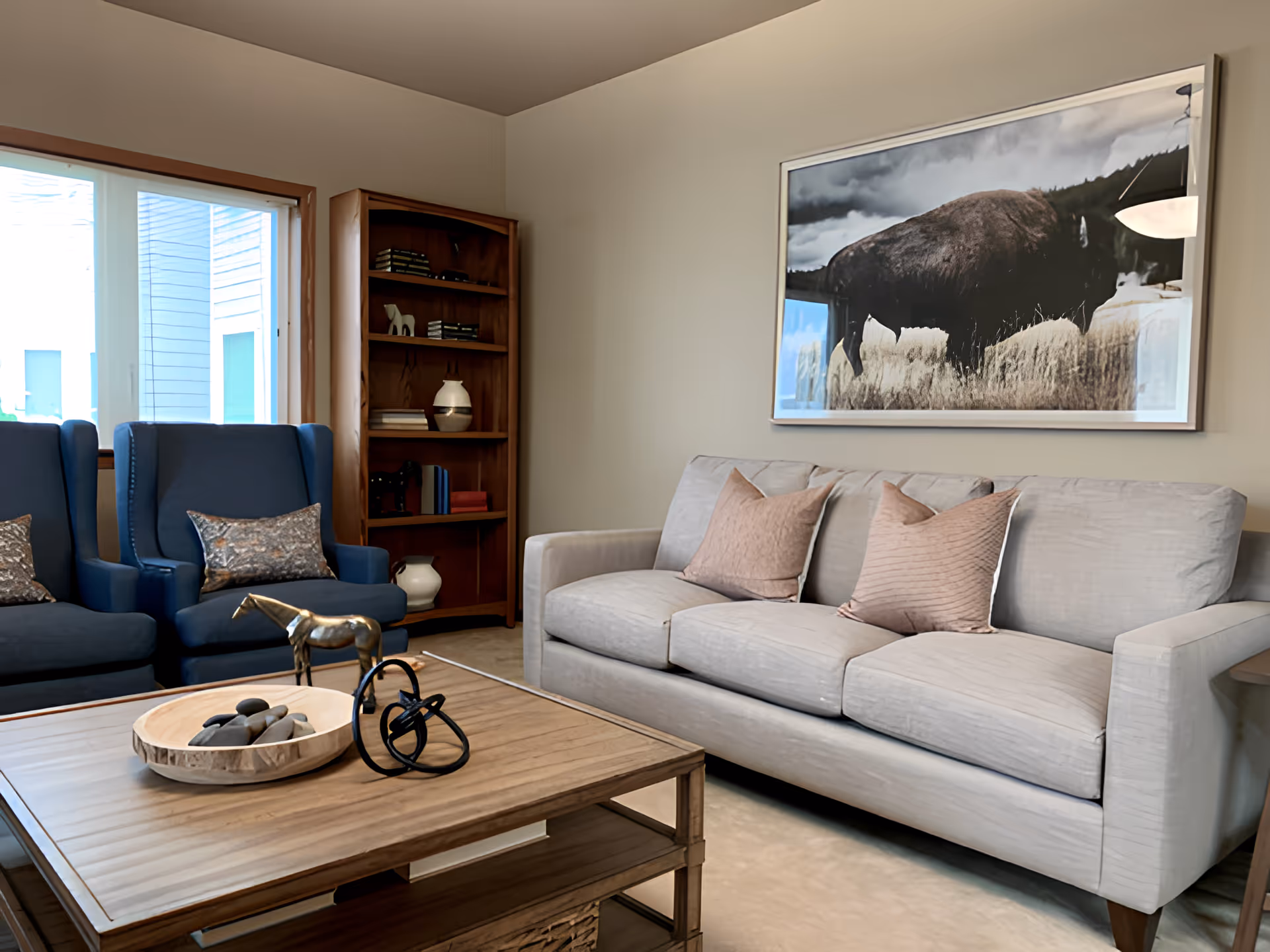 A cozy living room with a light gray sofa adorned with two pinkish throw pillows, two blue armchairs with patterned cushions, a wooden coffee table with decorative items including a bowl of stones and a small horse figurine, a wooden bookshelf with books and vases, and a large framed picture of a bison on the wall.