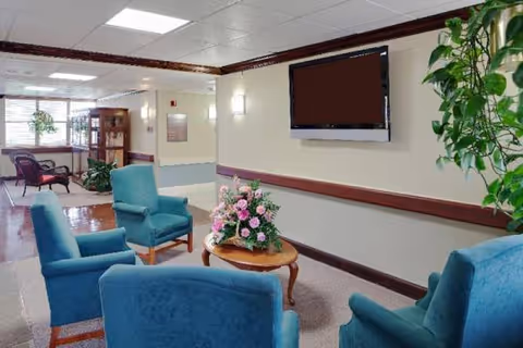 Bright senior living lounge with blue chairs, flowers, and wall-mounted TV.