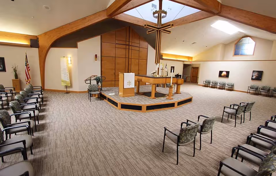 Spacious chapel interior with a central altar and large wooden cross, rows of chairs, and a vaulted wood-beamed ceiling.
