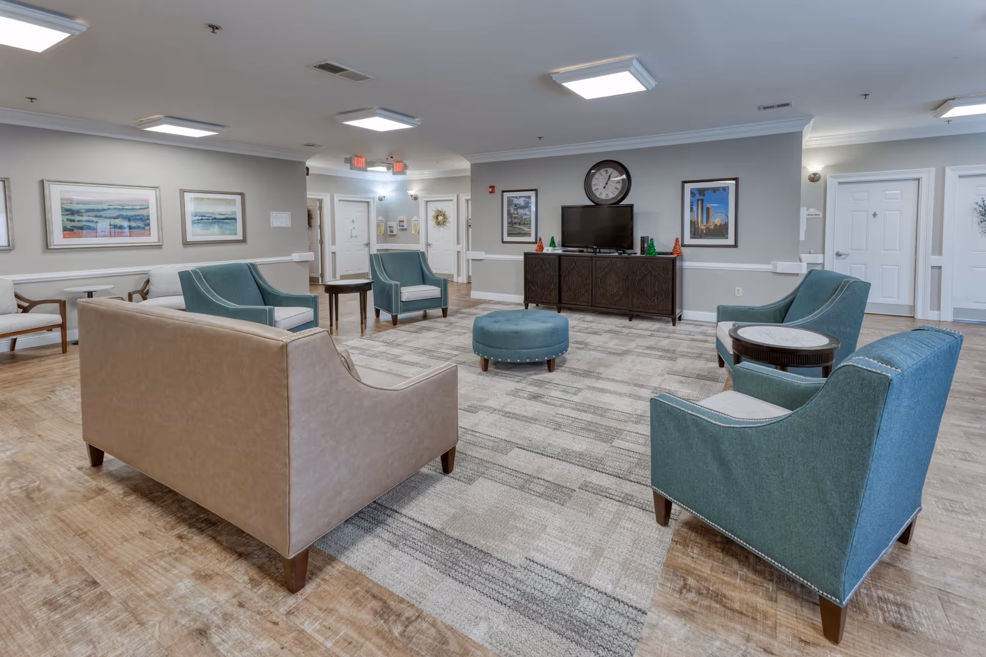 A spacious and well-lit common living area in a senior living facility featuring a beige sofa, four teal armchairs, a round blue ottoman, and a dark wooden TV cabinet with a flat-screen television and a large wall clock above it. The room has light wooden flooring, a large patterned area rug, framed artwork on the walls, and multiple white doors leading to other rooms.