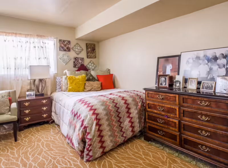 A cozy bedroom with a bed covered in a patterned comforter and multiple colorful pillows. Next to the bed is a wooden nightstand with a lamp and a framed photo. On the right side of the room is a wooden dresser with several framed photos on top. A window with sheer curtains allows natural light into the room, and a cushioned chair is placed near the nightstand.