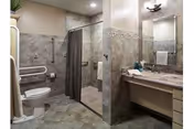 A spacious bathroom featuring a toilet with grab bars, a walk-in shower with a curtain, and a vanity with a large mirror and sink. The walls and floor are tiled in neutral tones, and there is a towel hanging near the shower.