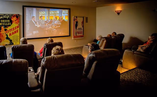 Several seniors seated in reclining chairs watch a movie projected on a screen in a dim community theater room.