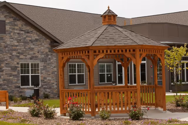 A wooden gazebo on landscaped grounds in front of a stone-and-siding building.