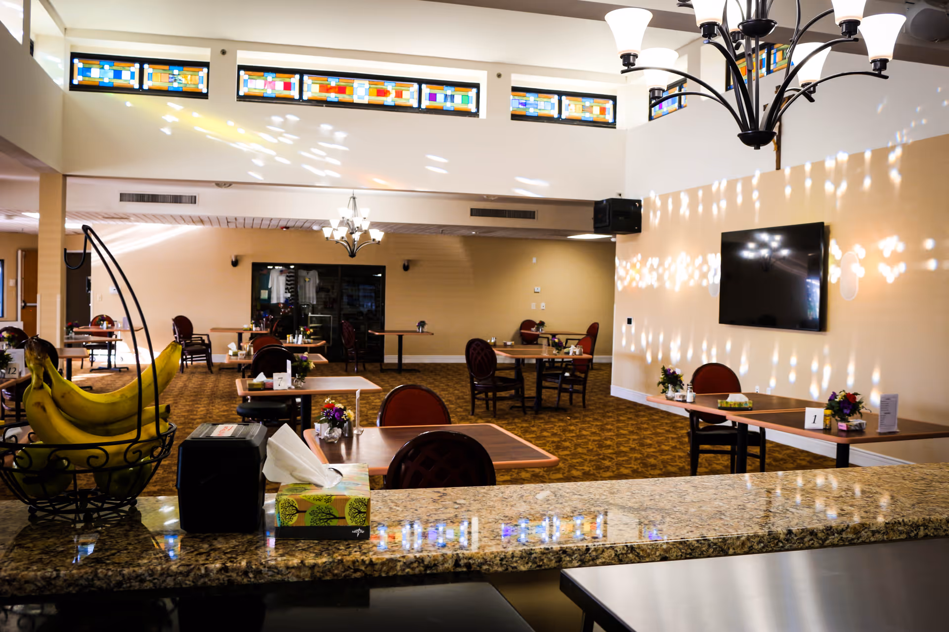 Interior view of a dining area in Villa St. Francis Catholic Care Center with several tables and chairs arranged neatly. The room features stained glass windows near the ceiling, a chandelier, and a wall-mounted flat screen TV. A countertop in the foreground holds a basket of bananas, a tissue box, and a napkin dispenser.
