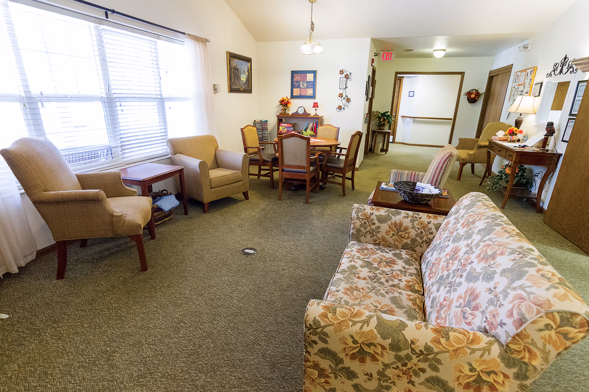 Communal lounge area with a floral sofa, armchairs, a small dining table, and large windows letting in light.