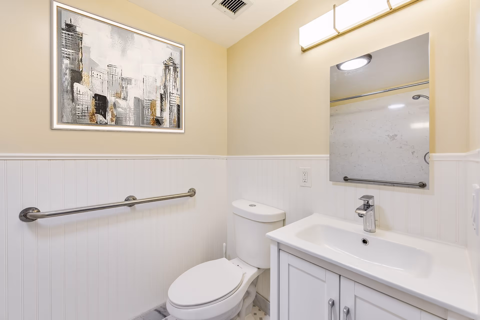 A clean bathroom with a white toilet and a white vanity with a sink. Above the sink is a rectangular mirror and a modern light fixture. The walls are painted beige on the upper half and white wainscoting on the lower half. A metal grab bar is mounted on the wall next to the toilet. There is a framed abstract cityscape painting hanging on the wall.
