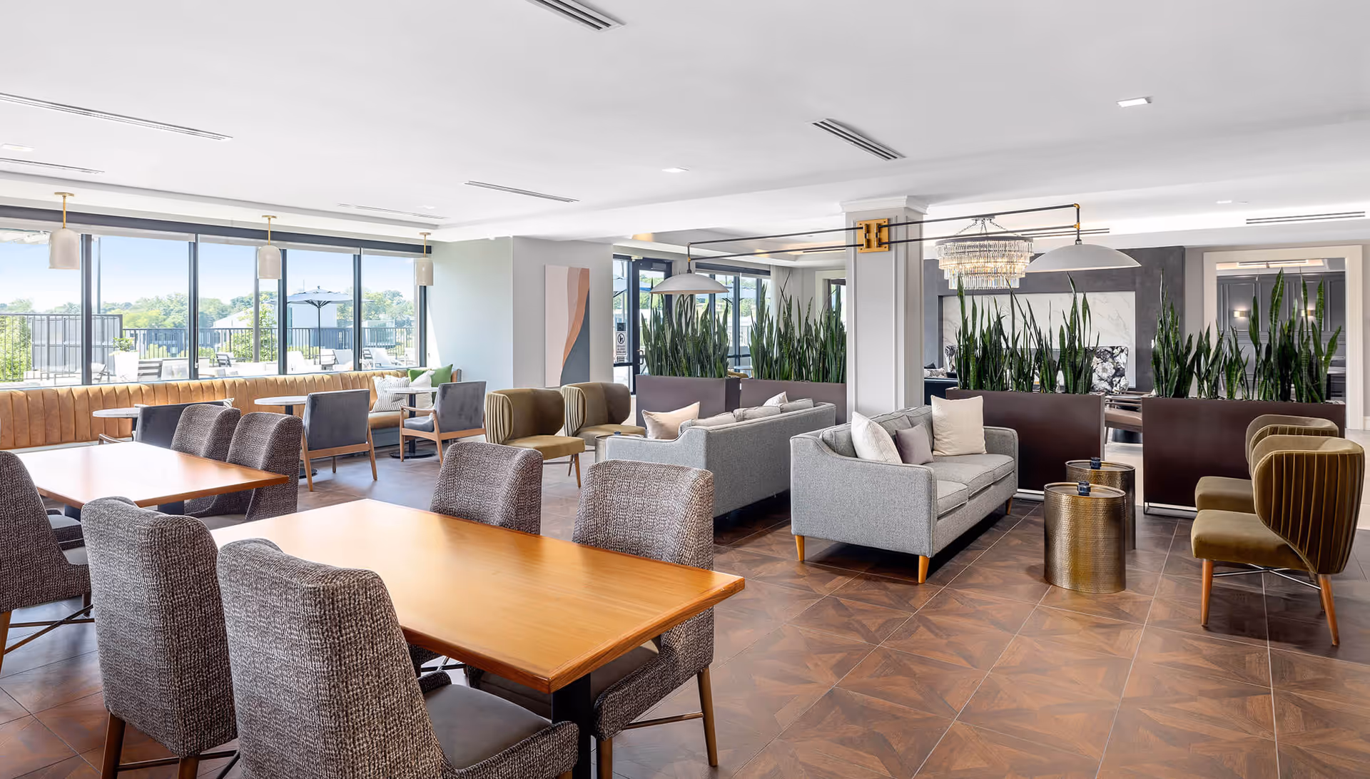 A spacious and modern common area in Avidor Omaha, featuring multiple seating arrangements including gray sofas, green velvet chairs, and wooden tables with upholstered chairs. Large windows allow natural light to fill the room, and tall green plants are used as room dividers. The floor has a patterned tile design and the ceiling has recessed lighting.