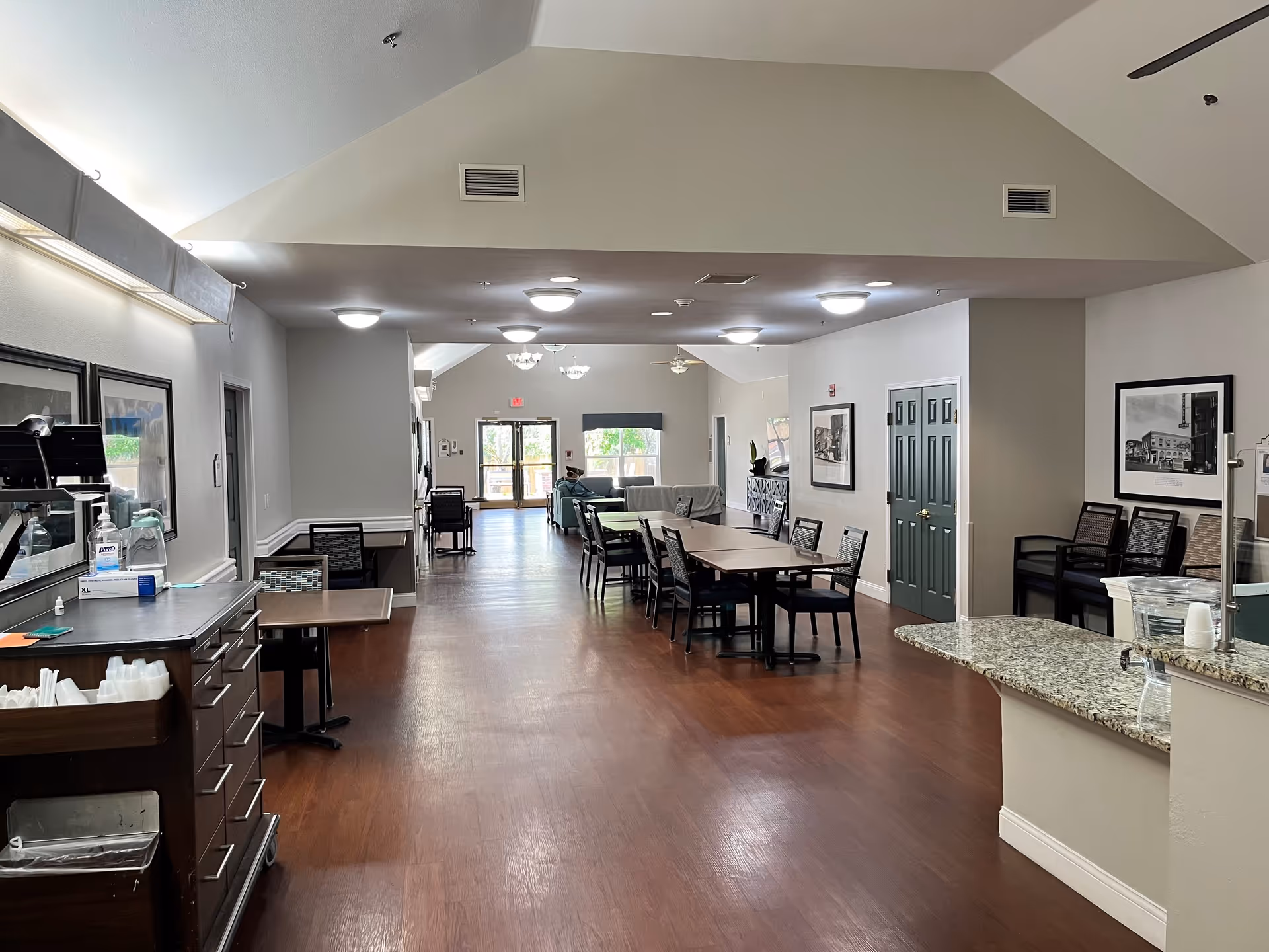 Interior view of a senior living facility common area with wooden flooring, tables and chairs arranged in the middle, a countertop with a water dispenser on the right, and framed pictures on the walls. The space is well-lit with ceiling lights and has a high vaulted ceiling. A person is seated in the far end near the windows and exit door.