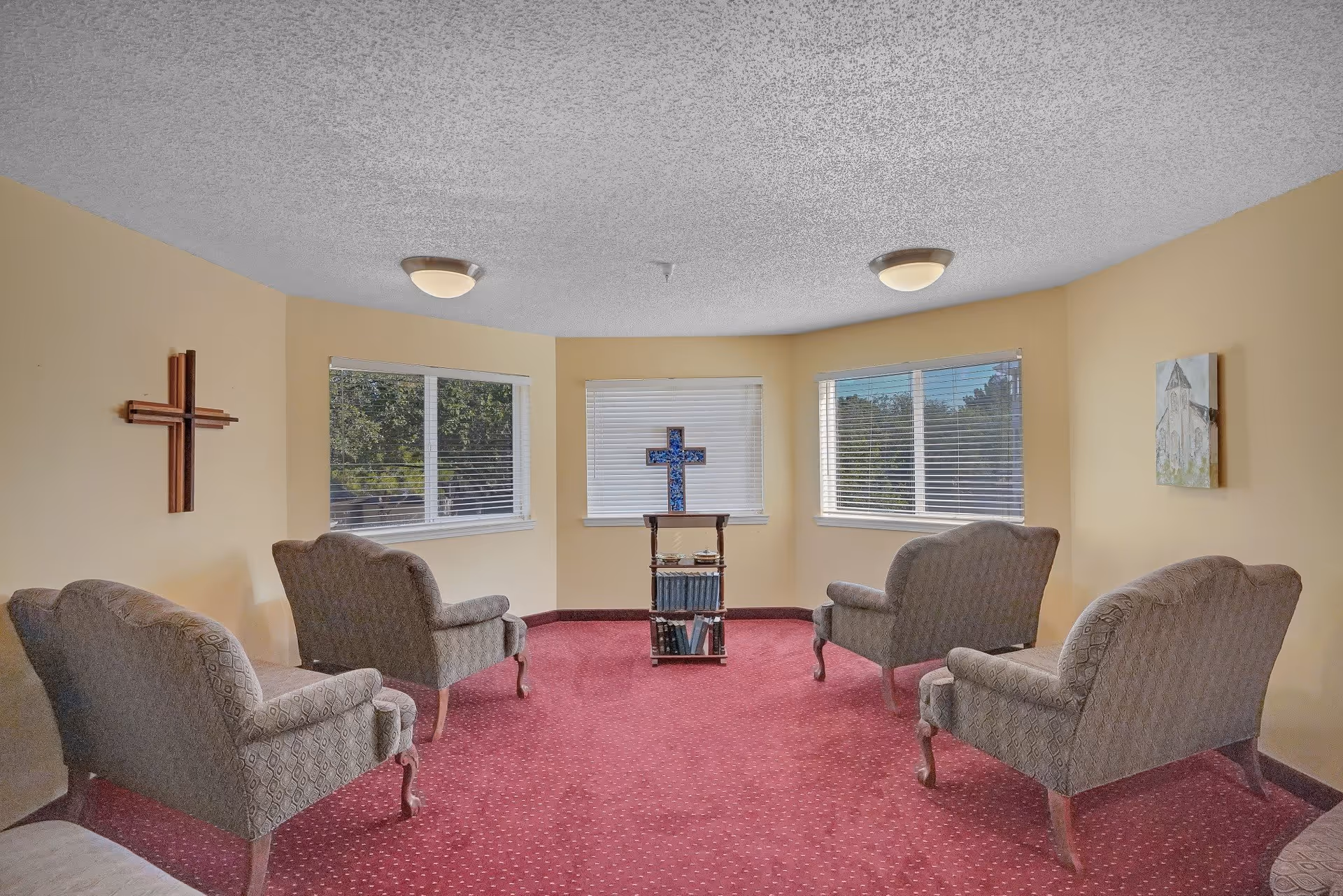 A small room with four upholstered armchairs arranged in a semi-circle on a red carpet. The walls are painted light yellow, and there are three windows with white blinds letting in natural light. A wooden cross is mounted on the left wall, and a small stand with books and a decorative cross is positioned in front of the center window. A framed picture hangs on the right wall.