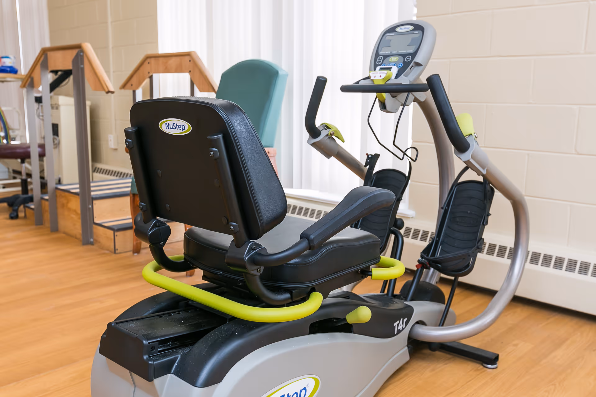 A NuStep T4r recumbent cross trainer exercise machine in a room with wooden floors and beige walls, next to a green chair and wooden handrails with steps.