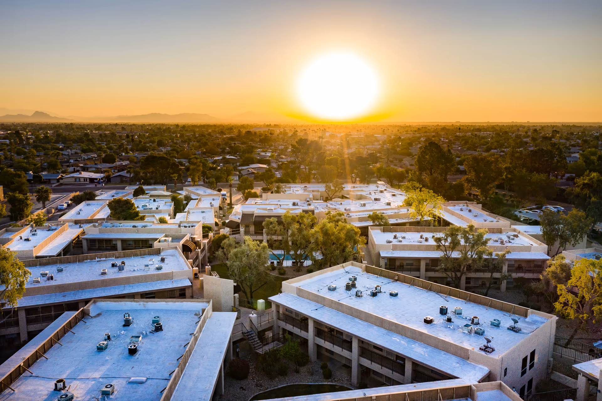 Aerial view of Scottsdale Village Square senior living facility at sunset, showing multiple flat-roofed buildings surrounded by trees and a suburban neighborhood extending into the distance with mountains on the horizon.