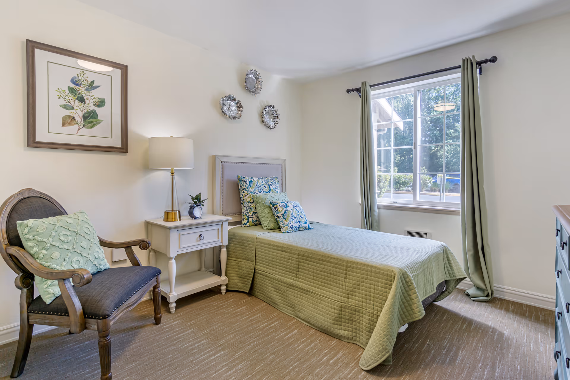 A neatly arranged bedroom with a single bed covered in a green quilt and multiple decorative pillows. Next to the bed is a white nightstand with a lamp, a small plant, and an alarm clock. A wooden chair with a green cushion is placed beside the nightstand. The room has a large window with green curtains, beige carpet flooring, and light-colored walls adorned with framed artwork and decorative wall pieces.