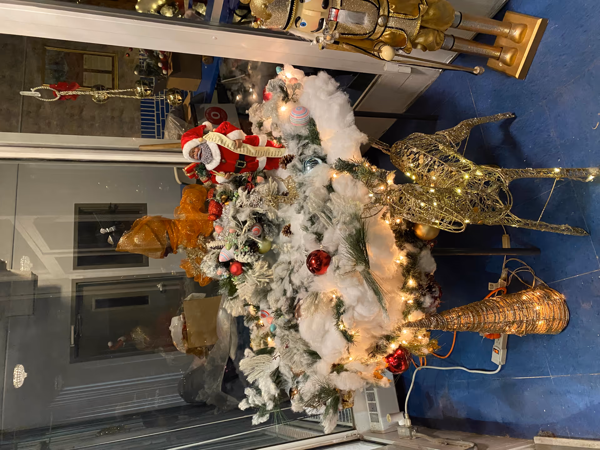 Indoor holiday display featuring a flocked, ornamented arrangement with lights, a Santa figure, and gold decorative reindeer near a glass window.