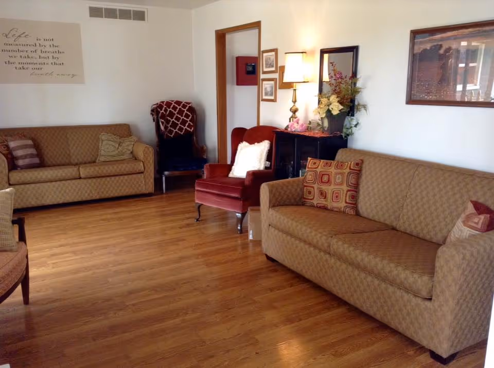 A cozy living room with two beige patterned sofas, a red armchair with a white pillow, a wooden floor, a side table with a lamp and flower arrangement, and framed pictures on the walls.