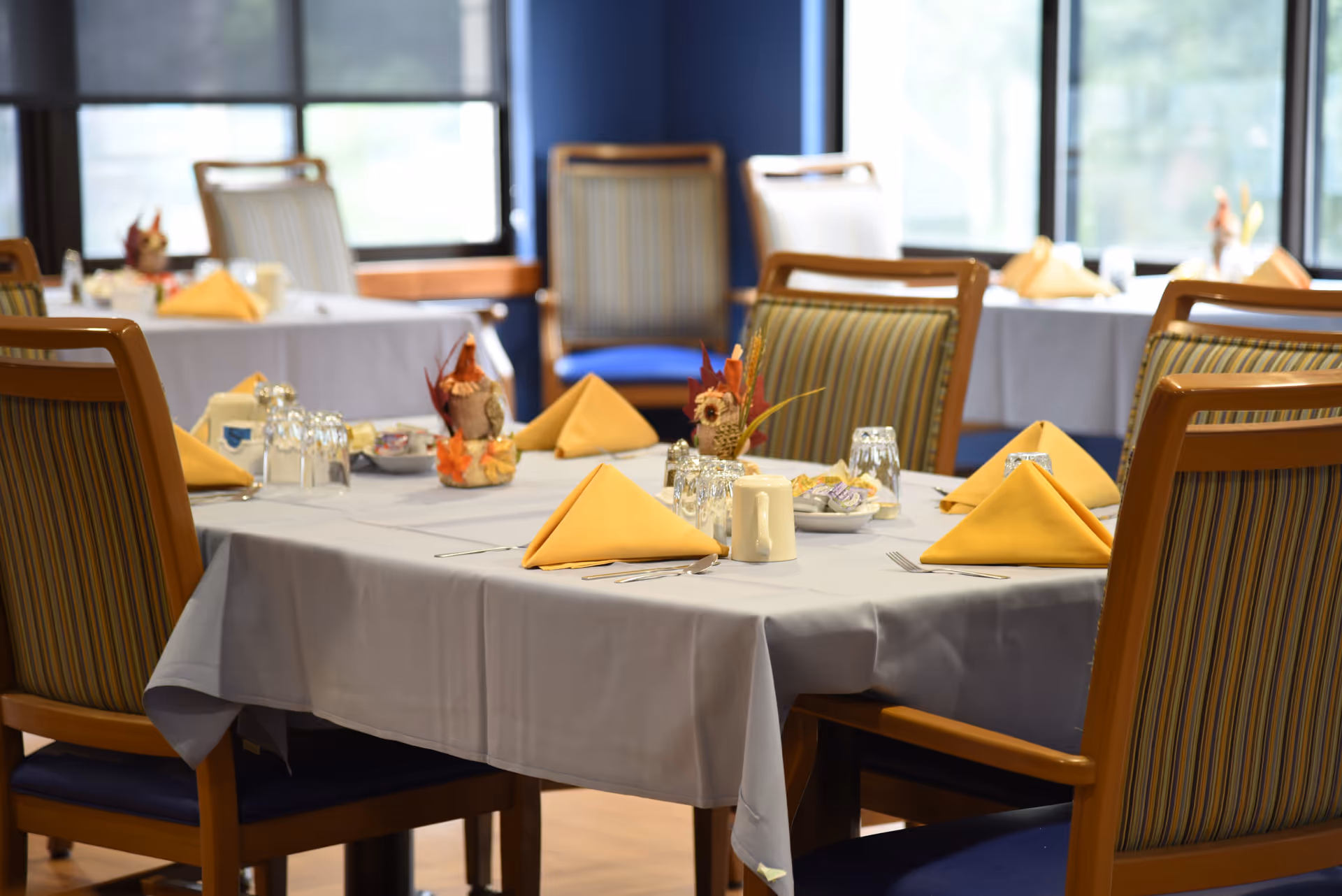 A dining room table set with a white tablecloth, yellow folded napkins, glassware, silverware, and small decorative centerpieces. The chairs around the table have wooden frames with striped upholstery. Large windows in the background let in natural light.
