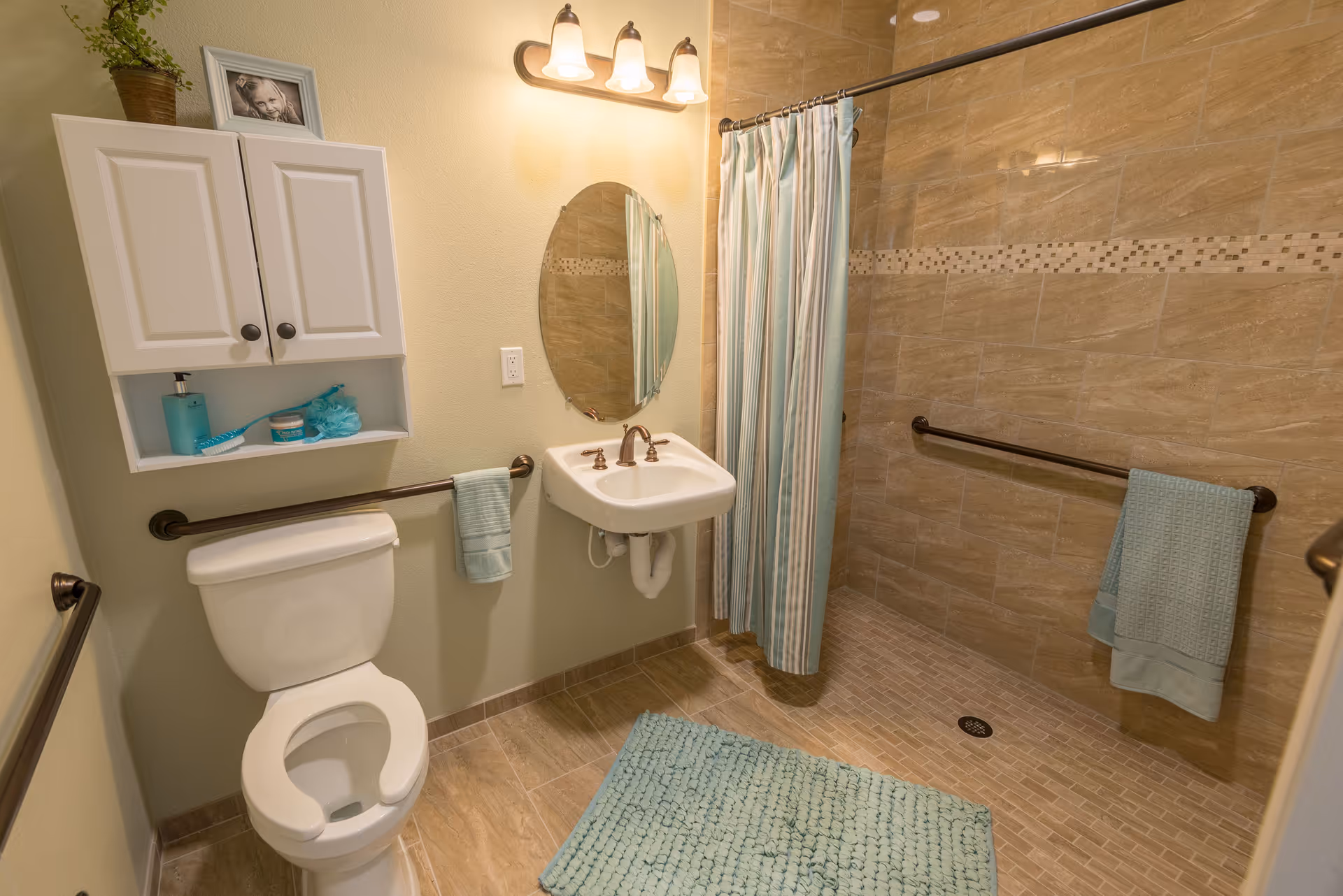A bathroom with beige tiled floor and walls, featuring a white toilet with a safety rail, a white wall-mounted sink with bronze faucet, an oval mirror above the sink, a shower area with a striped curtain, and matching blue towels and bath mat. There is a white cabinet mounted on the wall above the toilet with a small plant and a framed photo on top.