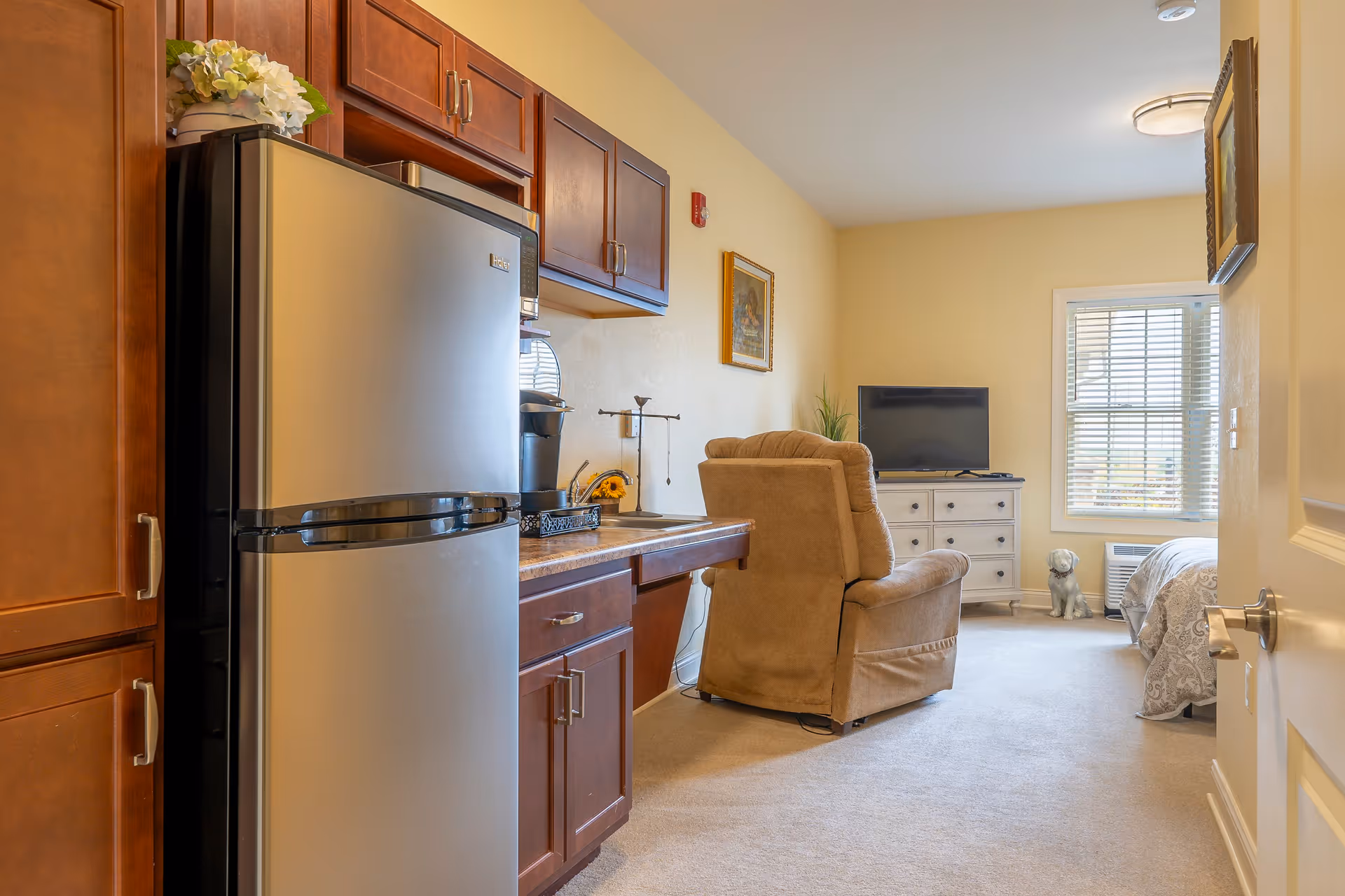 Interior view of a senior living facility room featuring a small kitchenette with a refrigerator, cabinets, and a coffee maker on the counter. The room also includes a beige recliner chair facing a flat-screen TV on a white dresser, a window with blinds, a bed partially visible on the right, and a small decorative dog statue near the window.