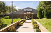 Exterior view of a single-story building with a peaked roof and a sign that reads 'aperion care' above the entrance. A paved walkway lined with small bushes leads to the glass double doors. The building is surrounded by green grass, trees, and shrubs under a clear blue sky.