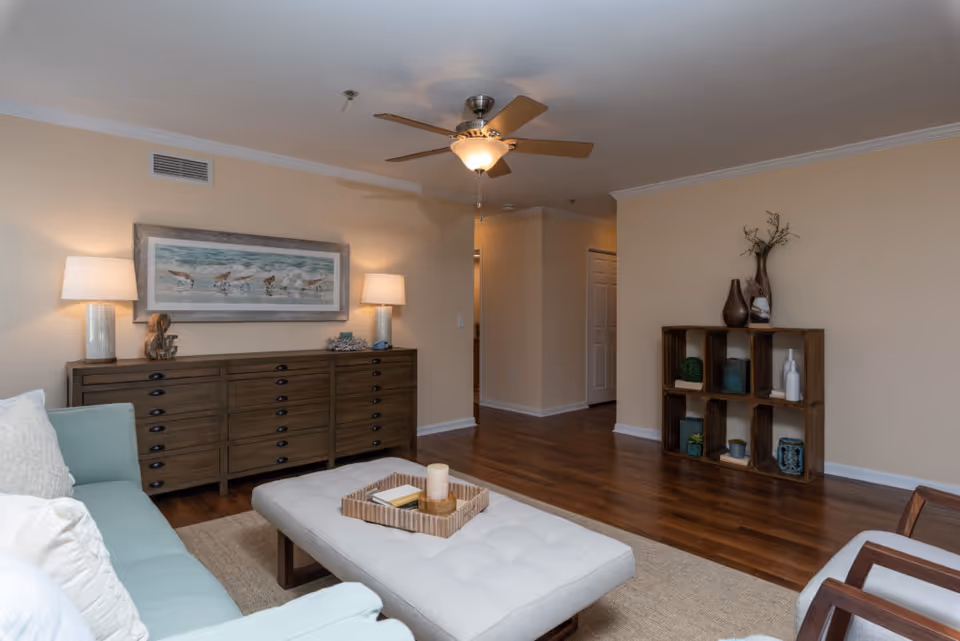 A cozy living room with a light blue sofa, a tufted ottoman with a tray holding a candle and a book, a wooden dresser with two lamps and a framed painting of birds above it, a wooden cube shelf with decorative items, and a ceiling fan with a light. The room has wooden flooring and beige walls.