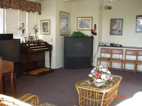 A living room with a large television set on a stand, an organ with decorative flowers on top, framed pictures on the walls, a glass-top wicker coffee table with a floral arrangement, and several chairs lined up against the wall.
