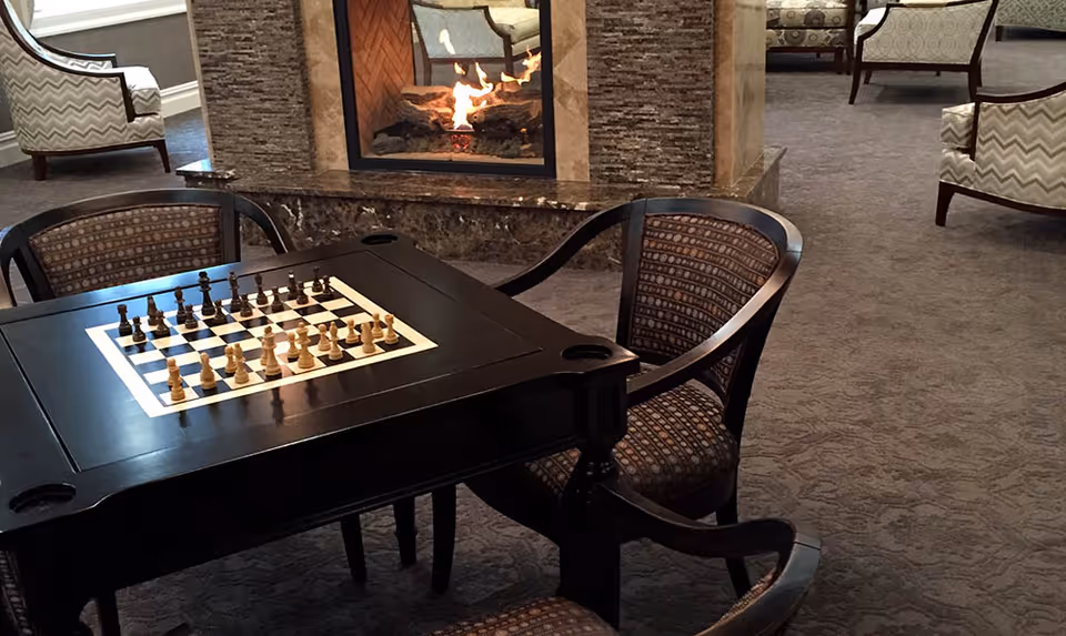 A cozy common area with a black chess table and four chairs arranged around it. A chess game is set up on the table. In the background, there is a lit fireplace with a stone surround and several upholstered armchairs placed on a carpeted floor.