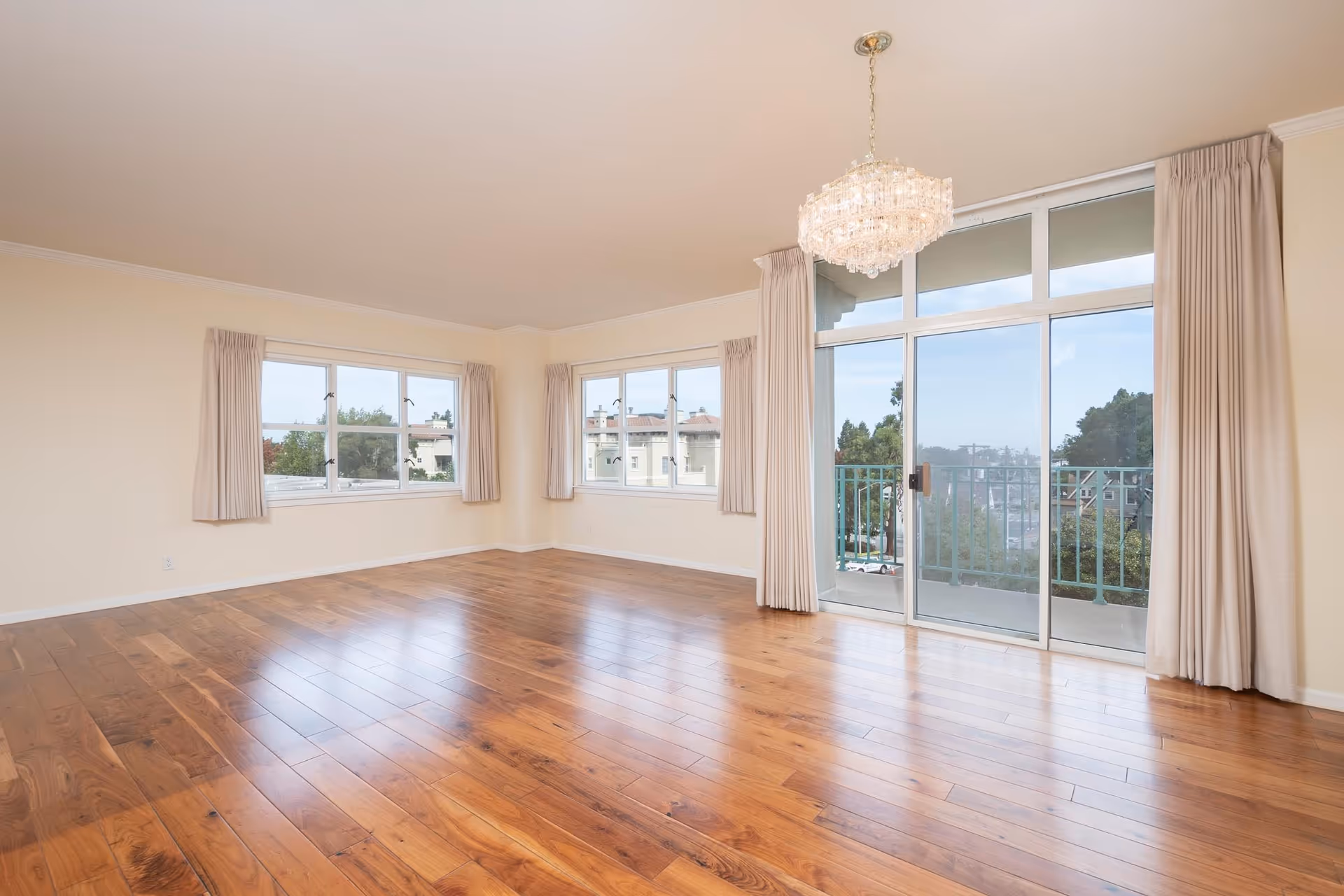 Empty room with polished wooden floor, cream-colored walls, multiple windows with beige curtains, a glass sliding door leading to a balcony, and a crystal chandelier hanging from the ceiling.