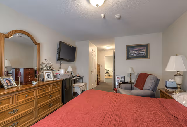 A bedroom with a red bedspread on the bed, a wooden dresser with a large mirror and framed photos, a wall-mounted TV, a small desk with a chair, and a gray armchair with a red throw blanket. The room has beige walls and carpeted floor, with a doorway leading to another room.