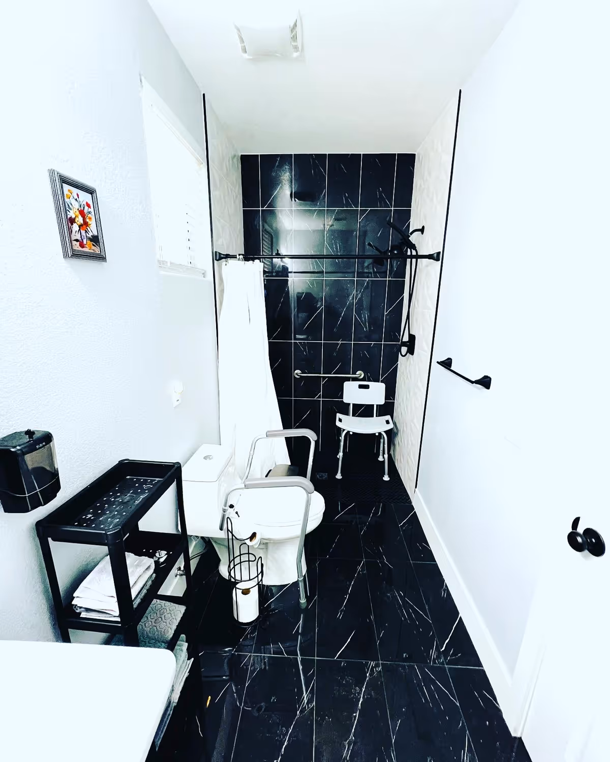 Accessible bathroom with a toilet fitted with safety rails, a shower chair, grab bars, and black marble-style tile floor and shower wall.