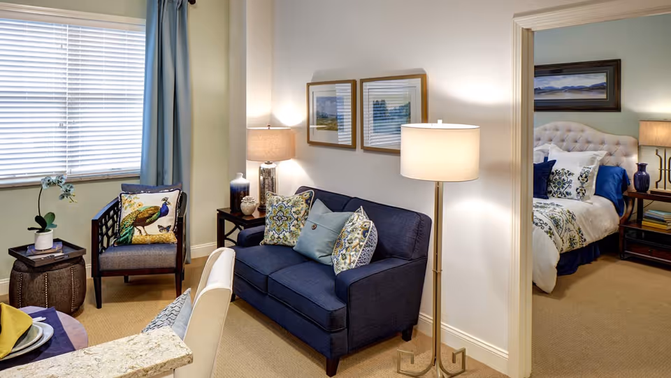 Small living area with a navy loveseat, patterned pillows, an armchair and floor lamp, opening into a furnished bedroom.