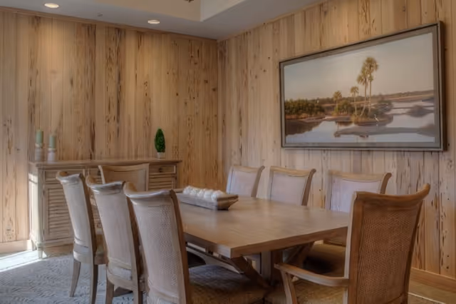 A dining room with a wooden table surrounded by eight upholstered chairs. The walls are covered with light wood paneling, and there is a sideboard with decorative items including candles and a small plant. A large framed landscape painting featuring palm trees and water hangs on the wall.