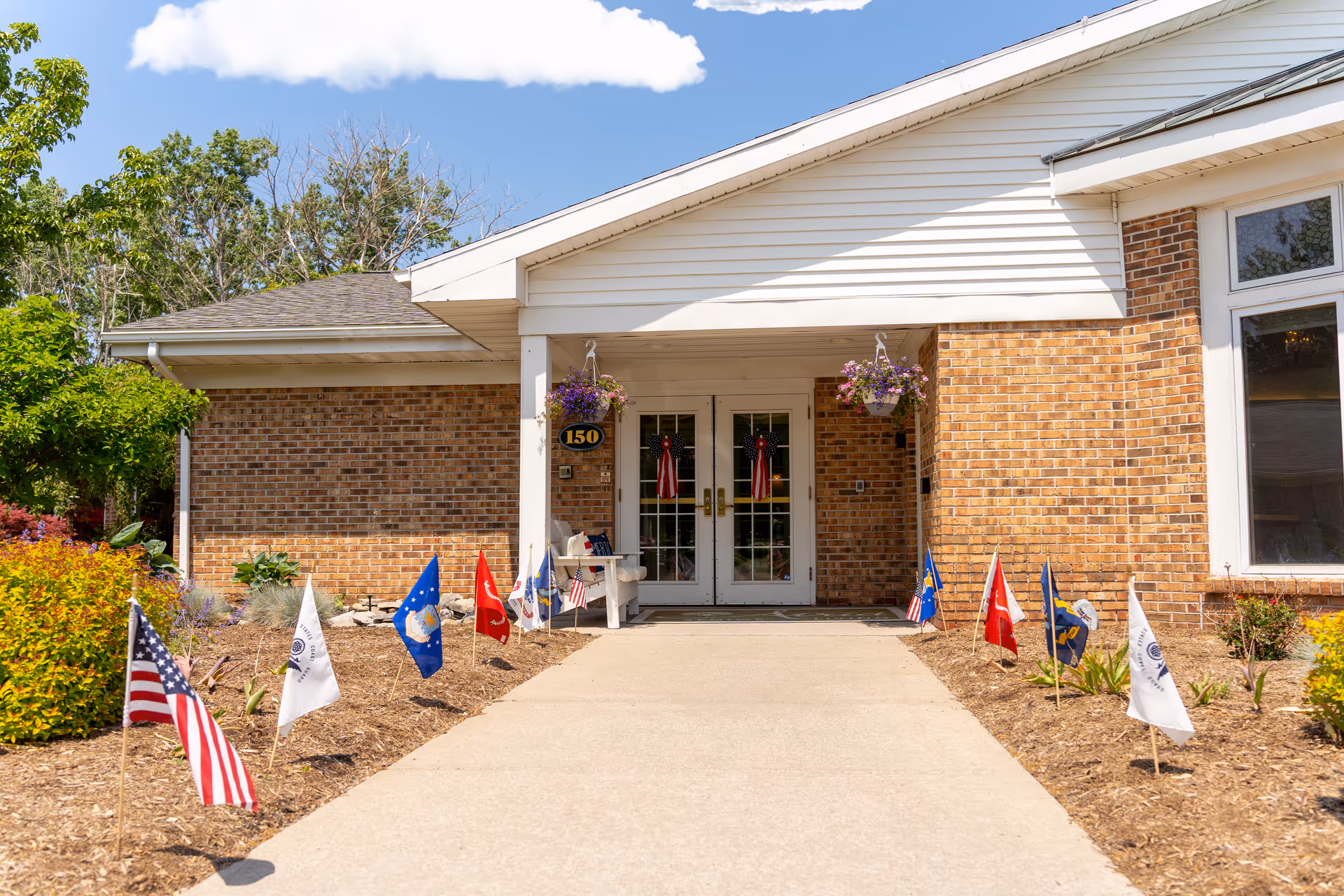 Entrance of a senior living community building with a brick exterior and white trim. A concrete walkway leads to double glass doors adorned with red, white, and blue decorations. Small flags line both sides of the walkway, and hanging flower baskets are visible near the entrance. The building number 150 is displayed on a plaque next to the door. Trees and shrubs surround the entrance under a partly cloudy blue sky.