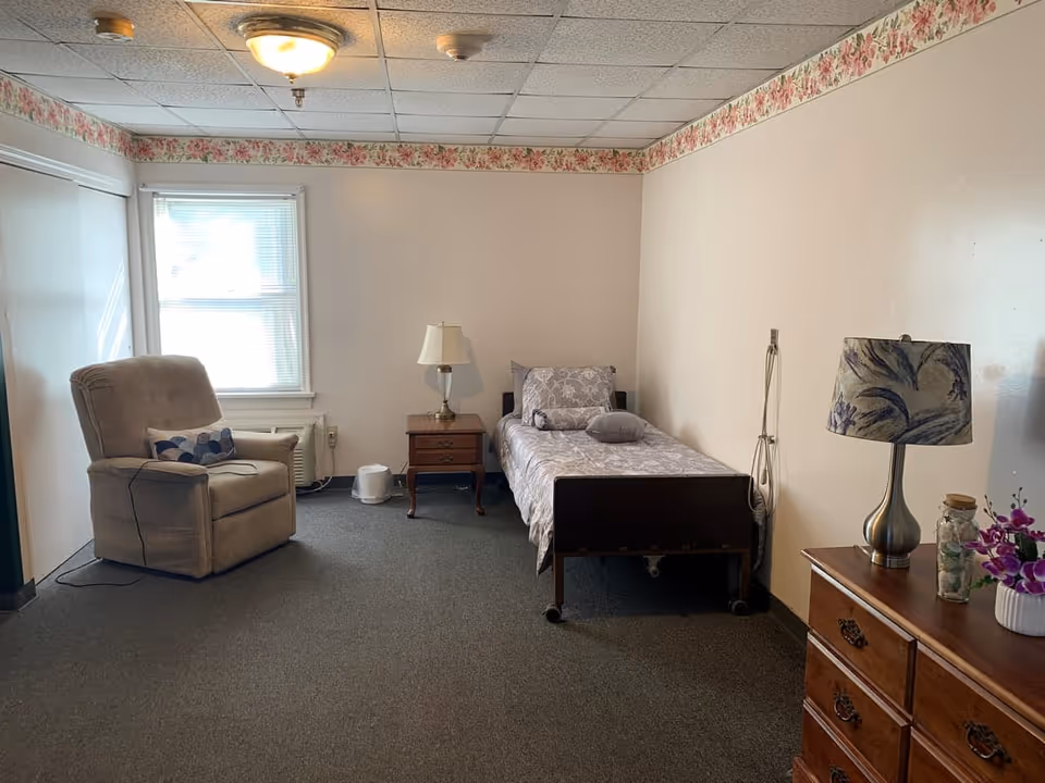 A simple senior living bedroom with a single bed covered in patterned bedding, a wooden nightstand with a lamp, a beige recliner chair with a decorative pillow, a window with blinds, and a wooden dresser with a floral lamp and a small vase of purple flowers. The room has a floral wallpaper border near the ceiling and gray carpeted floor.