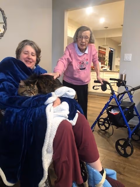 Two elderly women in a cozy indoor setting. One woman is sitting in a wheelchair wrapped in a blue blanket with a large cat resting on her lap. The other woman, wearing a pink sweater, is standing nearby using a blue walker. The background shows a kitchen area with warm lighting.