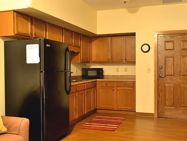 Interior view of a kitchen area with wooden cabinets, a black refrigerator, a microwave on the countertop, a wooden door, a small wall clock, and a red striped rug on the wooden floor.