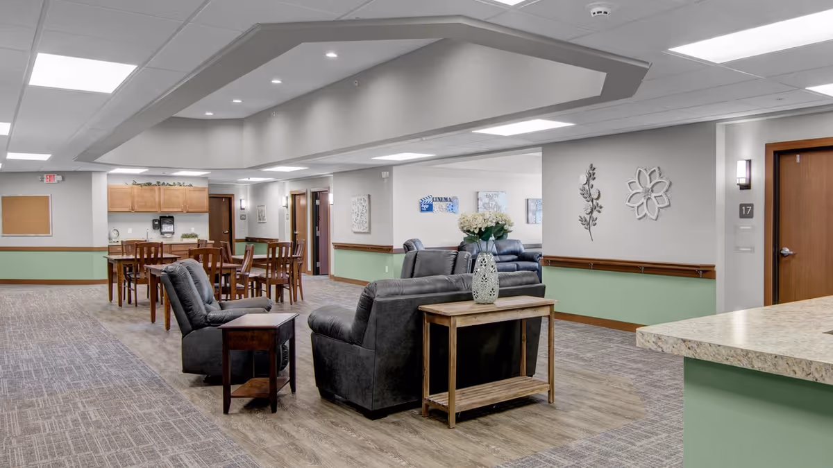 A spacious senior living common area with gray leather armchairs and sofas arranged around wooden side tables. In the background, there are wooden dining tables and chairs, light green and beige walls with decorative wall art, and a hallway with several doors. The ceiling has recessed lighting and a drop ceiling design.